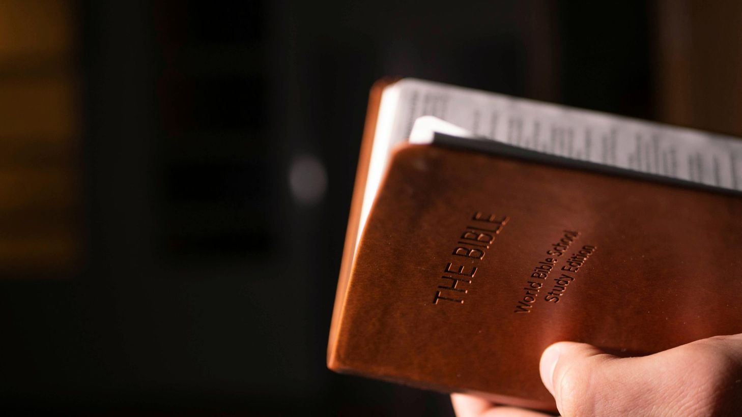 A close-up view of a hand holding a brown leather-bound Bible in a dimly lit room.