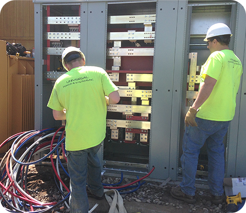 Two electricians in high-visibility yellow shirts working on the open electrical panels of a large outdoor cabinet.