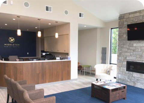 A waiting room with a wooden reception desk, brown armchairs, a white accent chair, and a stone fireplace with a TV.