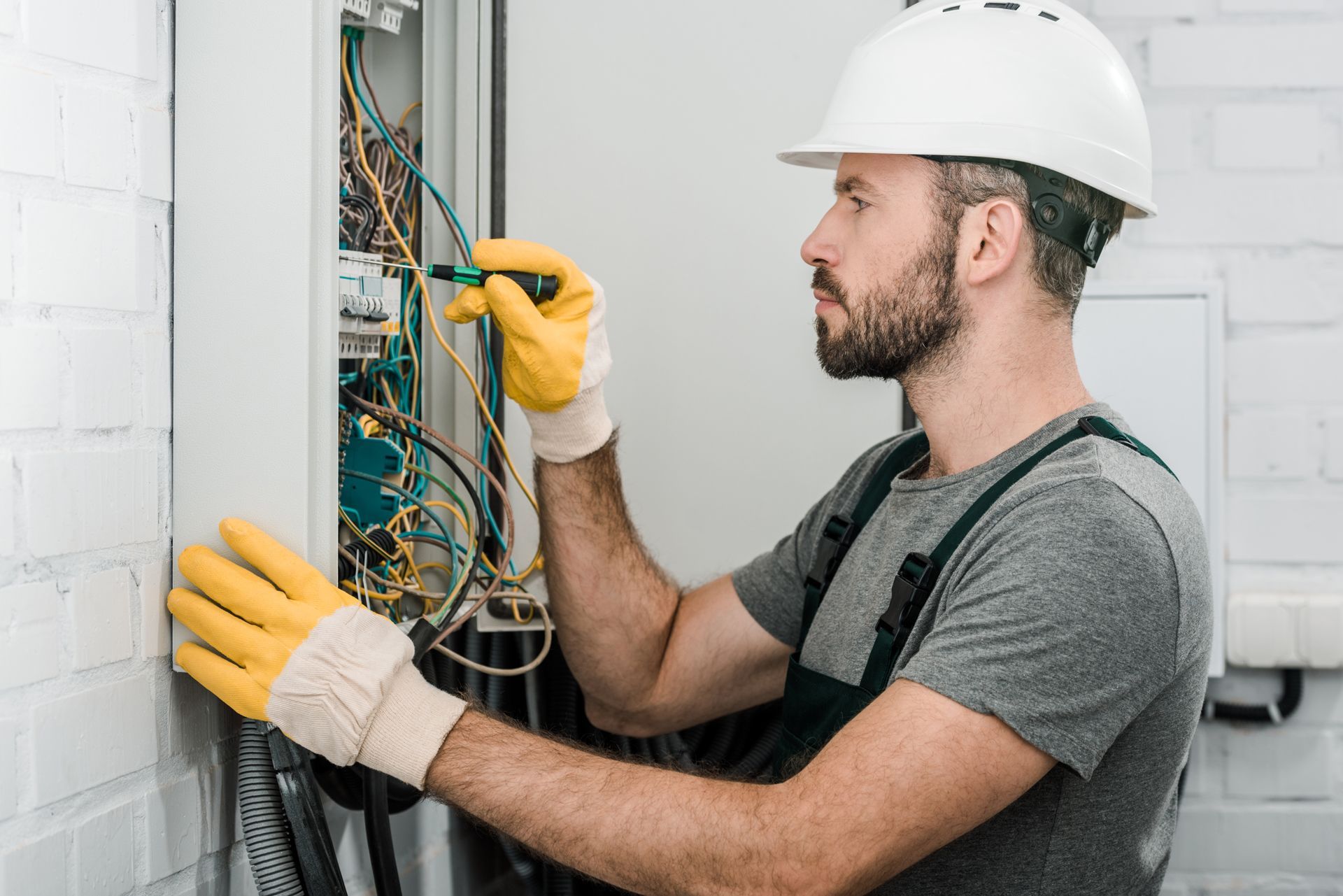 An electrician wearing a hard hat and protective gloves works on an open electrical panel against a white brick wall.