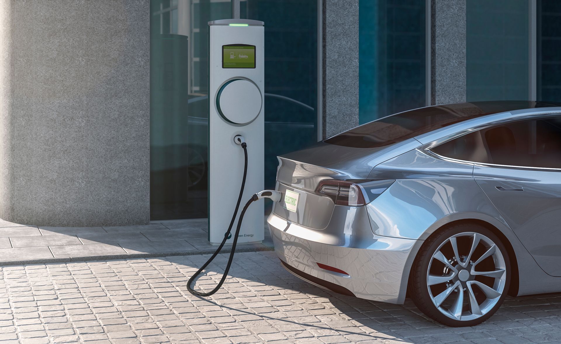 A silver electric car parked at an outdoor charging station with a black charging cable plugged into the vehicle.