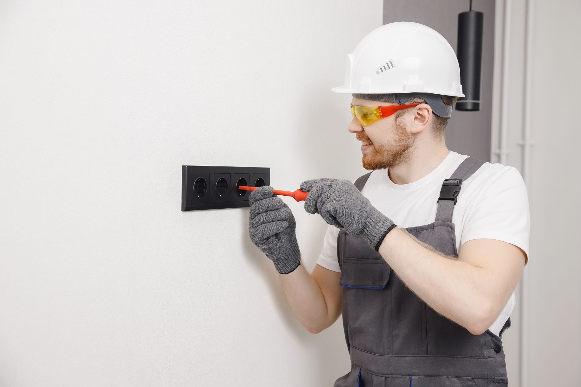 An electrician wearing a hard hat, safety glasses, and gloves uses a screwdriver on a black wall outlet.