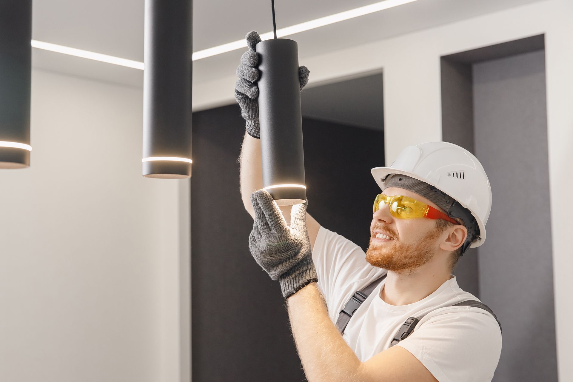 A worker in a hard hat and safety glasses installs a cylindrical pendant light fixture on a ceiling.