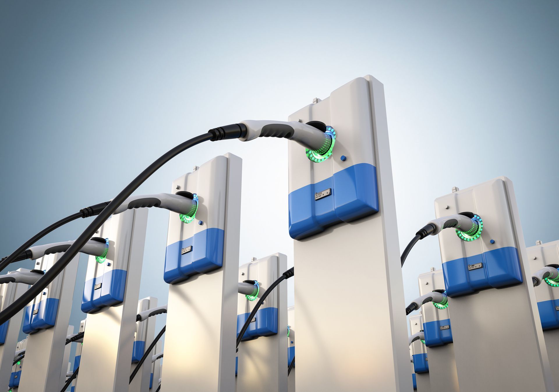 A row of electric vehicle charging stations with white and blue units, each with a black charging cable plugged in.