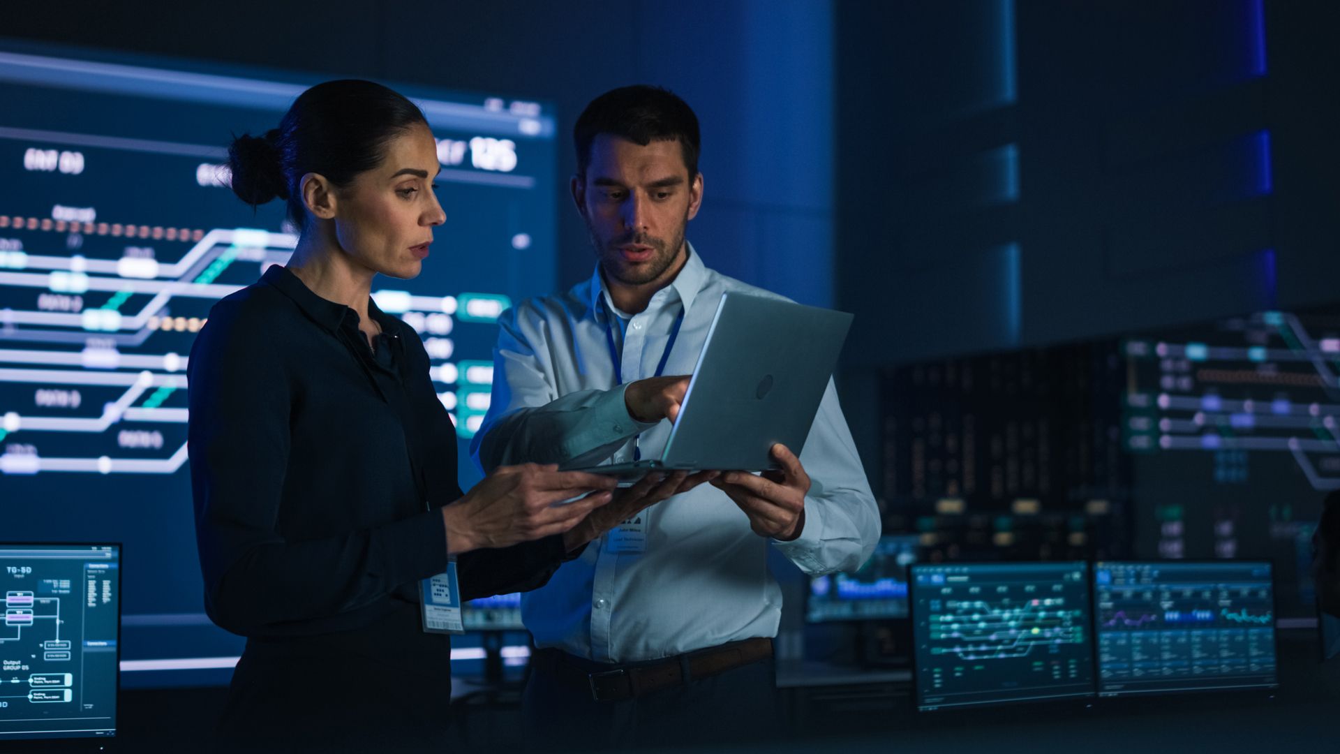 A woman and a man in a dimly lit control room review data on a laptop in front of large digital monitoring screens.