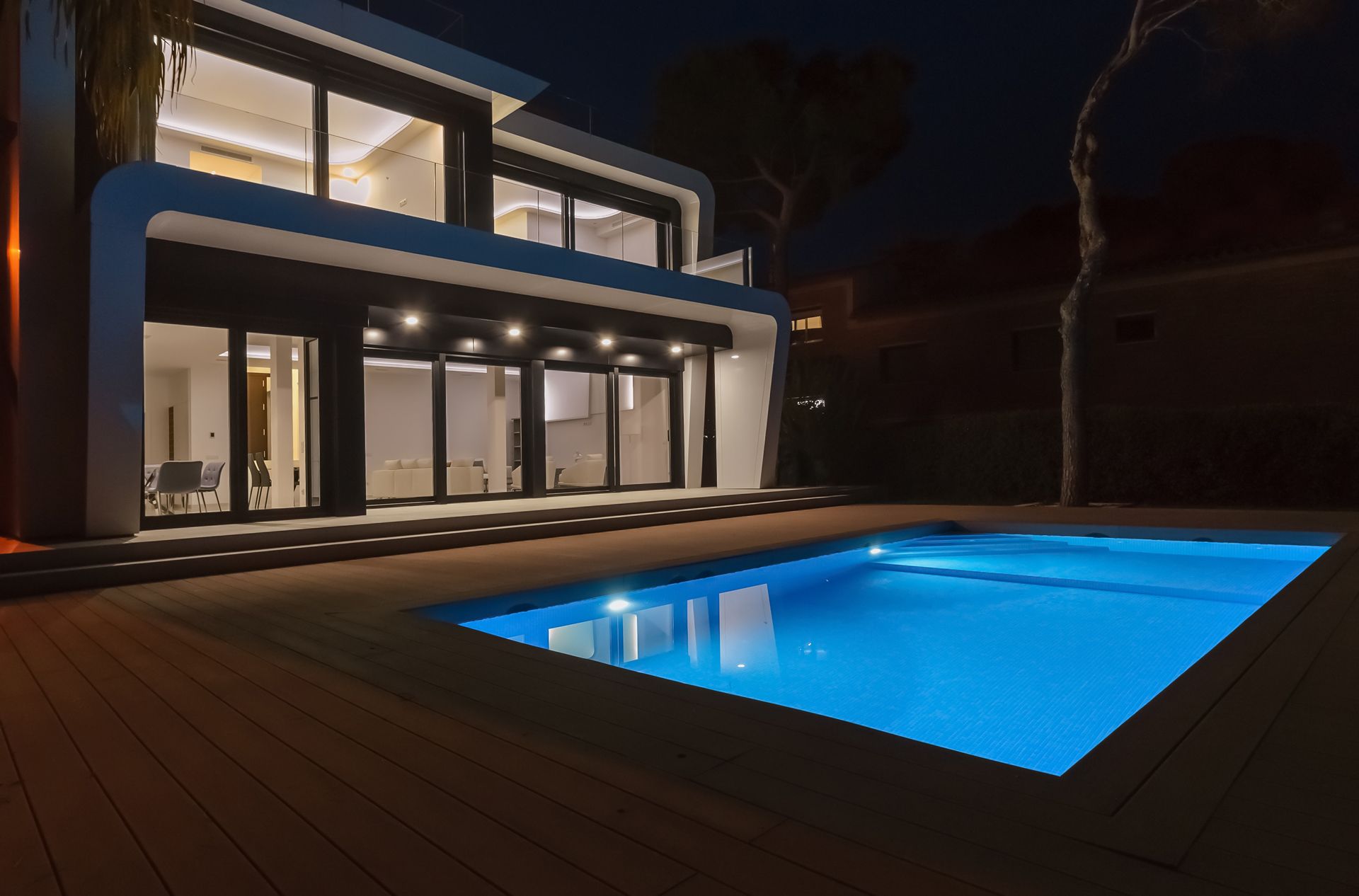 Modern two-story house with illuminated floor-to-ceiling windows and a blue-lit swimming pool at night.