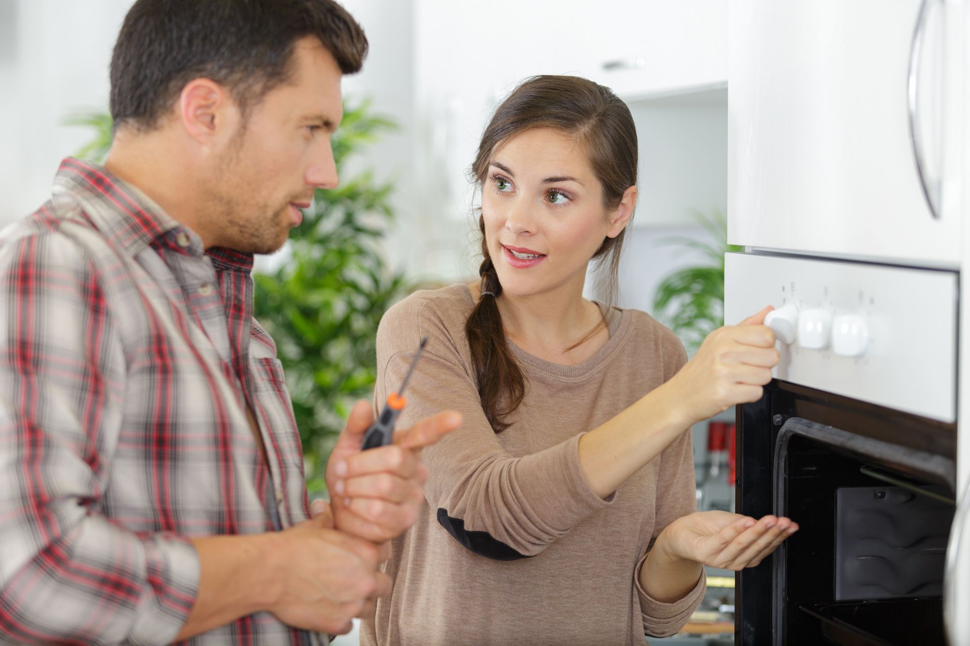 A person holds a screwdriver while talking to another person who is turning a dial on a wall-mounted oven.