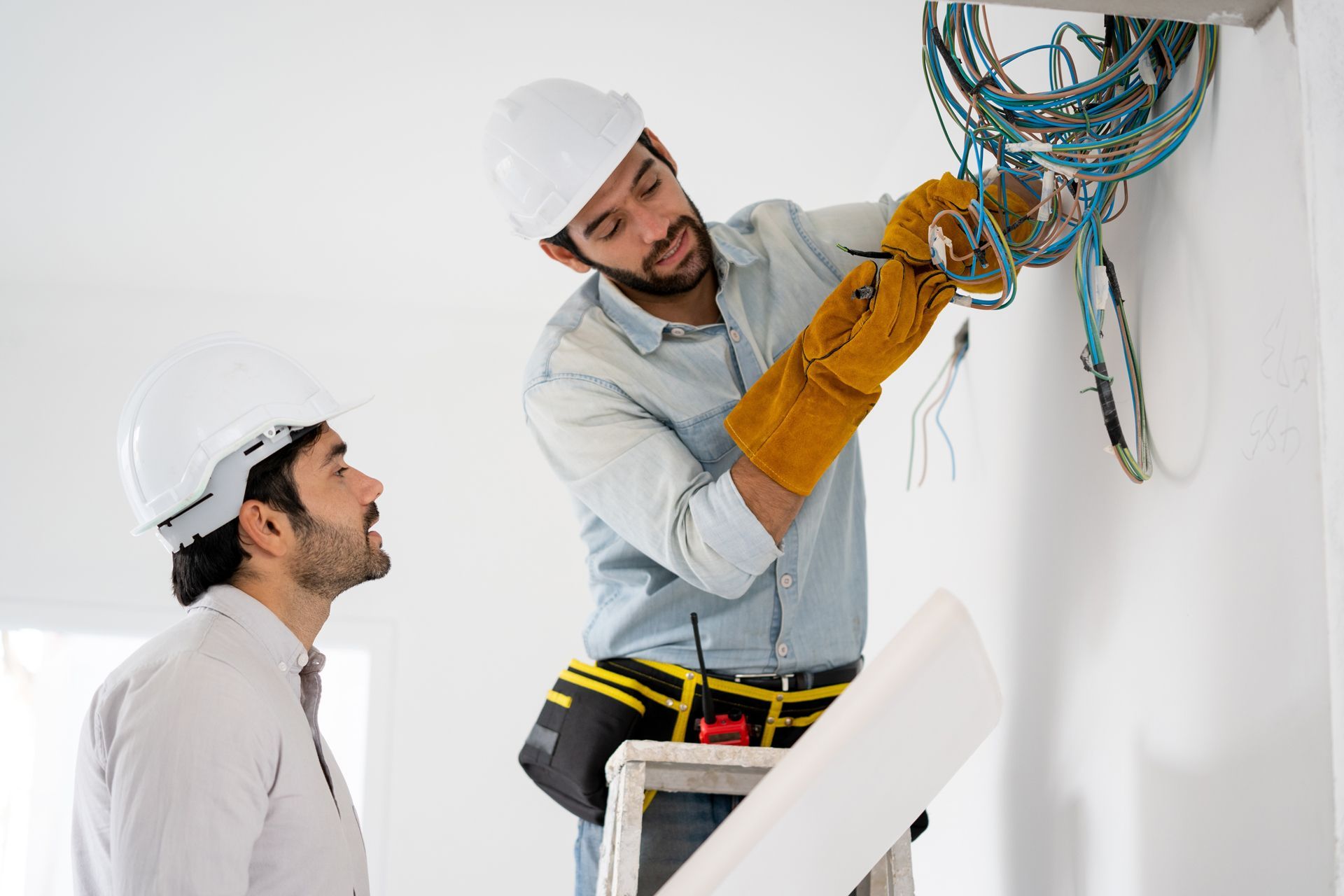 Two workers in hard hats and tool belts inspect electrical wiring exposed on a white interior wall.