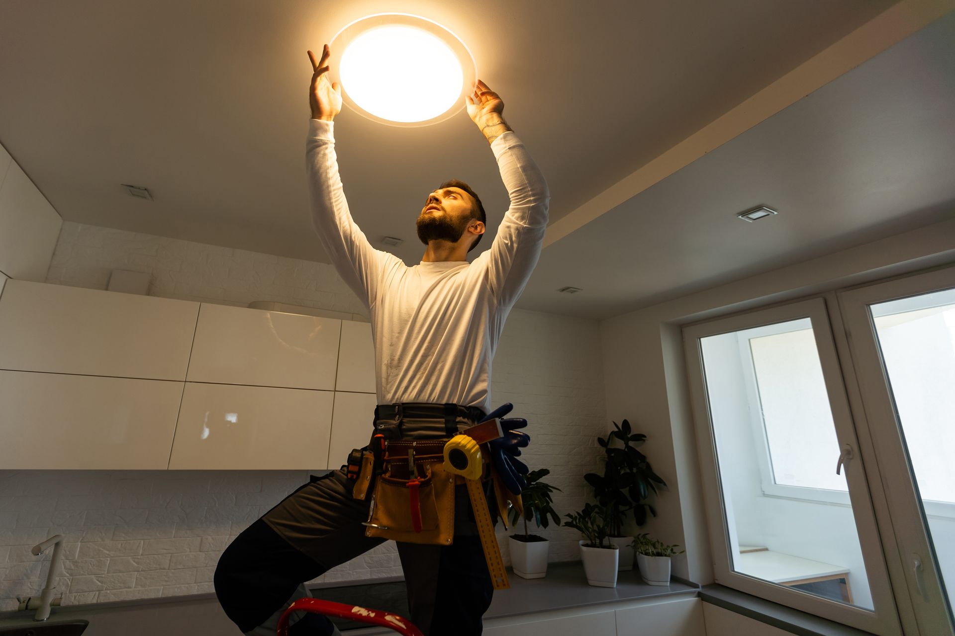 A person standing on a ladder, installing or adjusting a bright circular ceiling light in a kitchen.