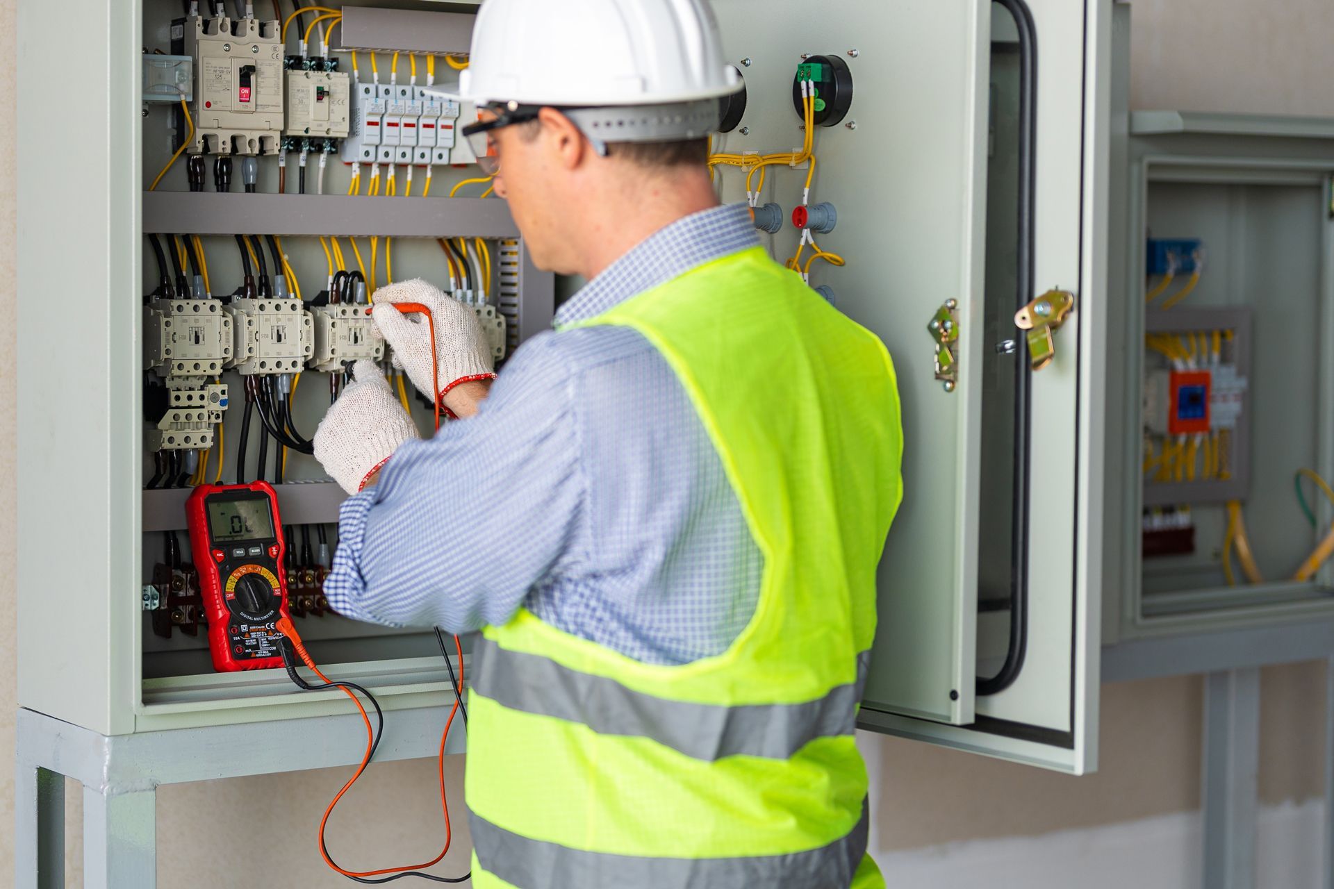 An electrician in a hard hat and safety vest tests electrical components in a control panel with a digital multimeter.
