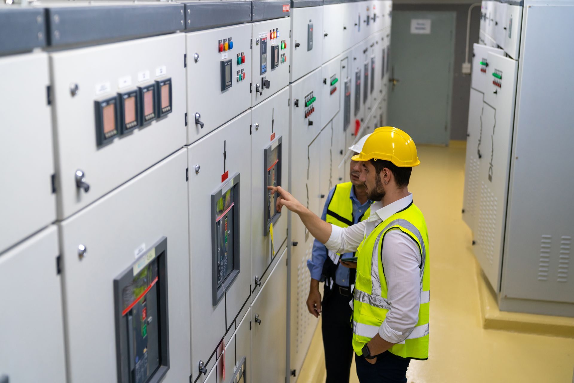 Two technicians in hard hats and safety vests inspect electrical control panels in a brightly lit industrial facility.
