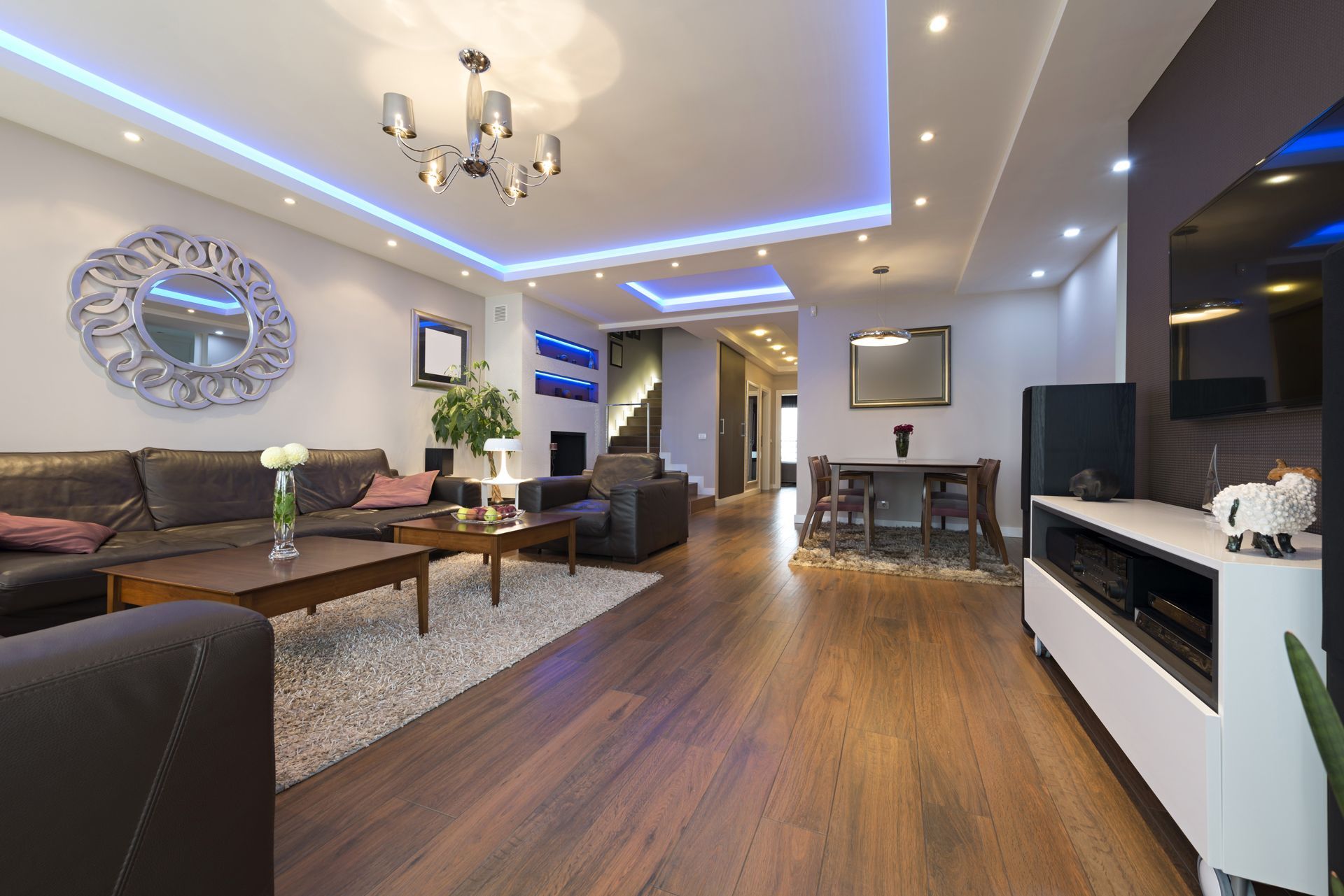A modern living room with wood floors, brown leather sofas, blue ceiling accent lighting, and a white TV console.