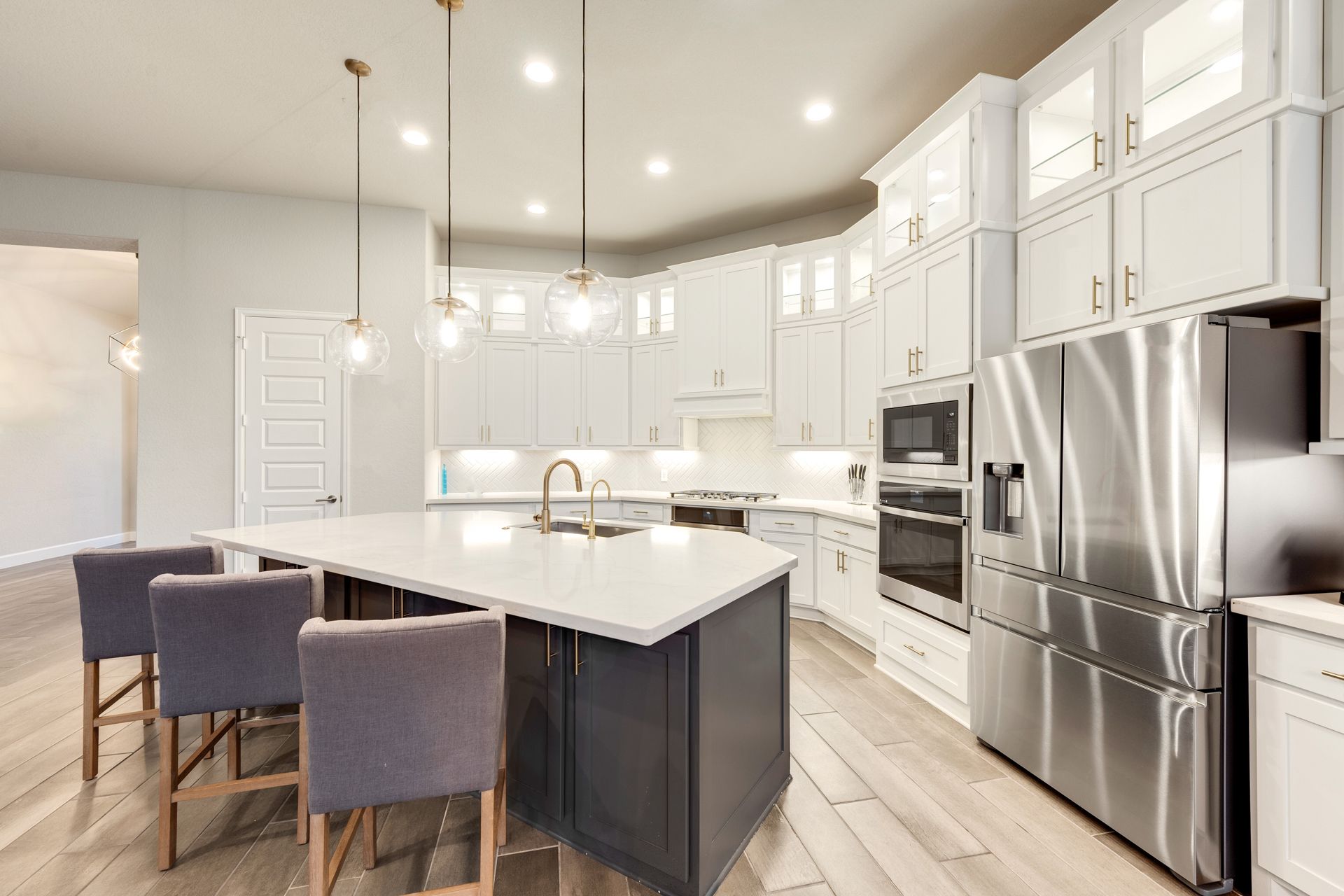 A modern kitchen with white cabinets, a dark island with three stools, stainless steel appliances, and wood floors.