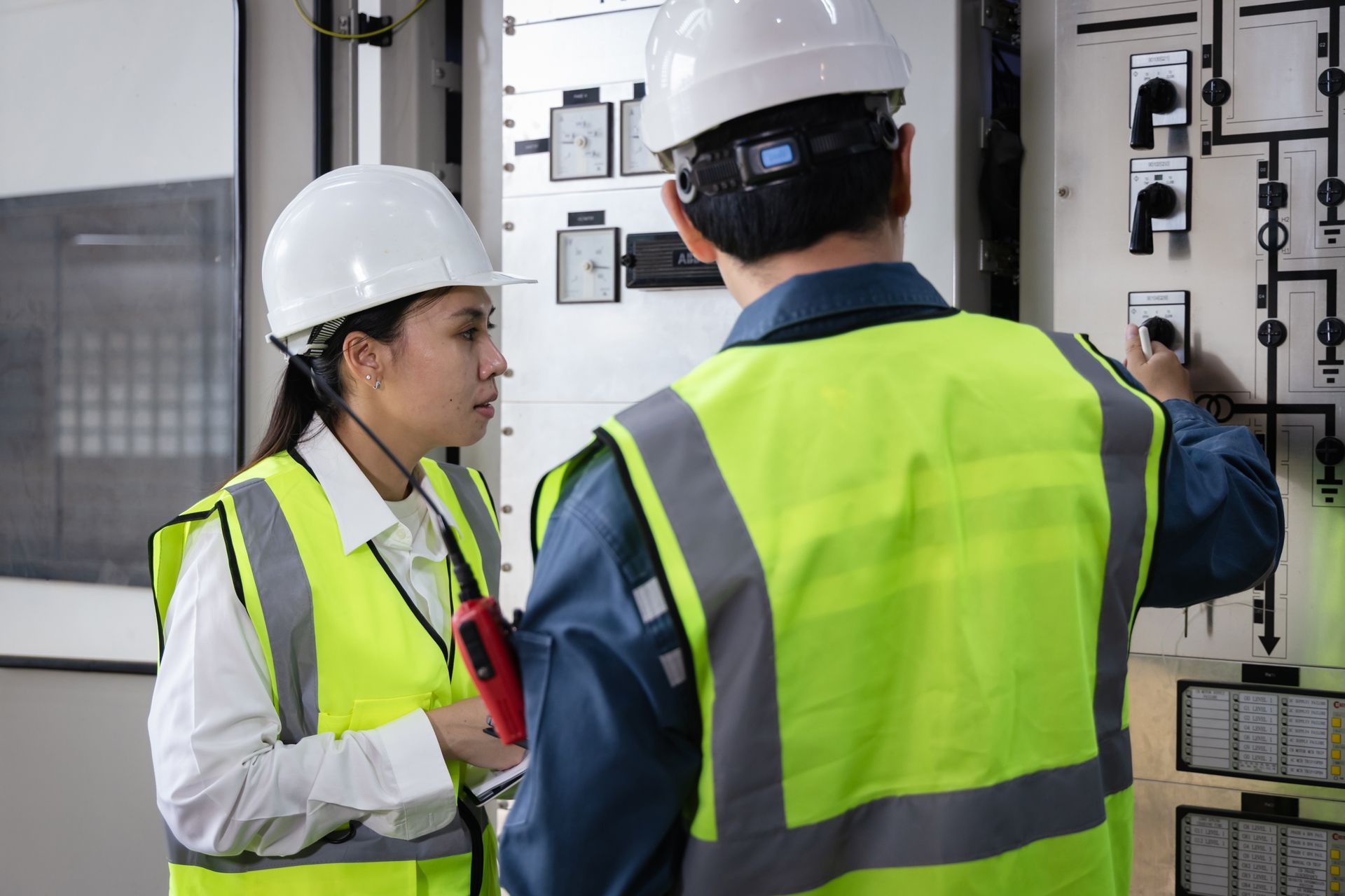 Two people in hard hats and high-visibility vests inspect electrical equipment in an industrial setting.