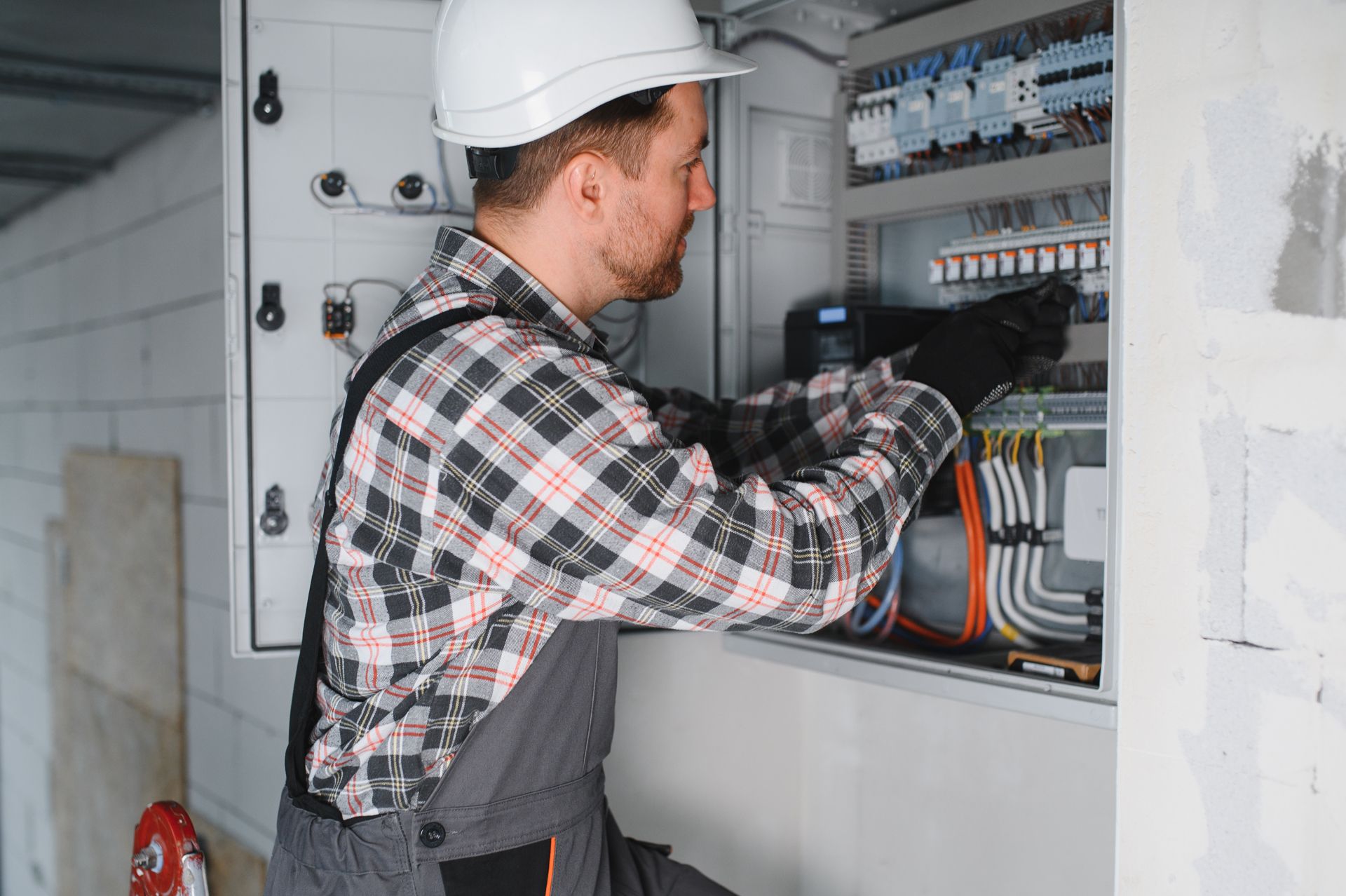 An electrician in a plaid shirt and hard hat installs or repairs wiring in an open electrical panel.
