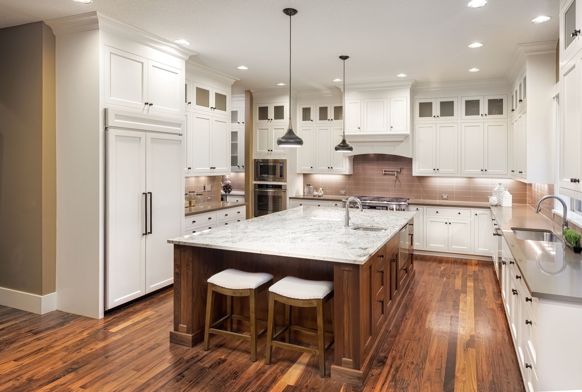 A bright kitchen with white cabinetry, a large marble-topped island, wooden floors, and two hanging pendant lights.