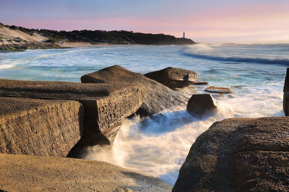 Waves Crashing On A Rocky Beach With A Lighthouse In The Distance — The Kitchen & Bathroom Doctor in Soldiers Point, NSW