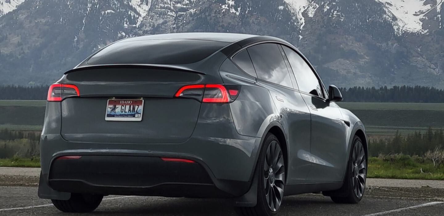 A white tesla model y is parked in a gravel lot in front of a building.