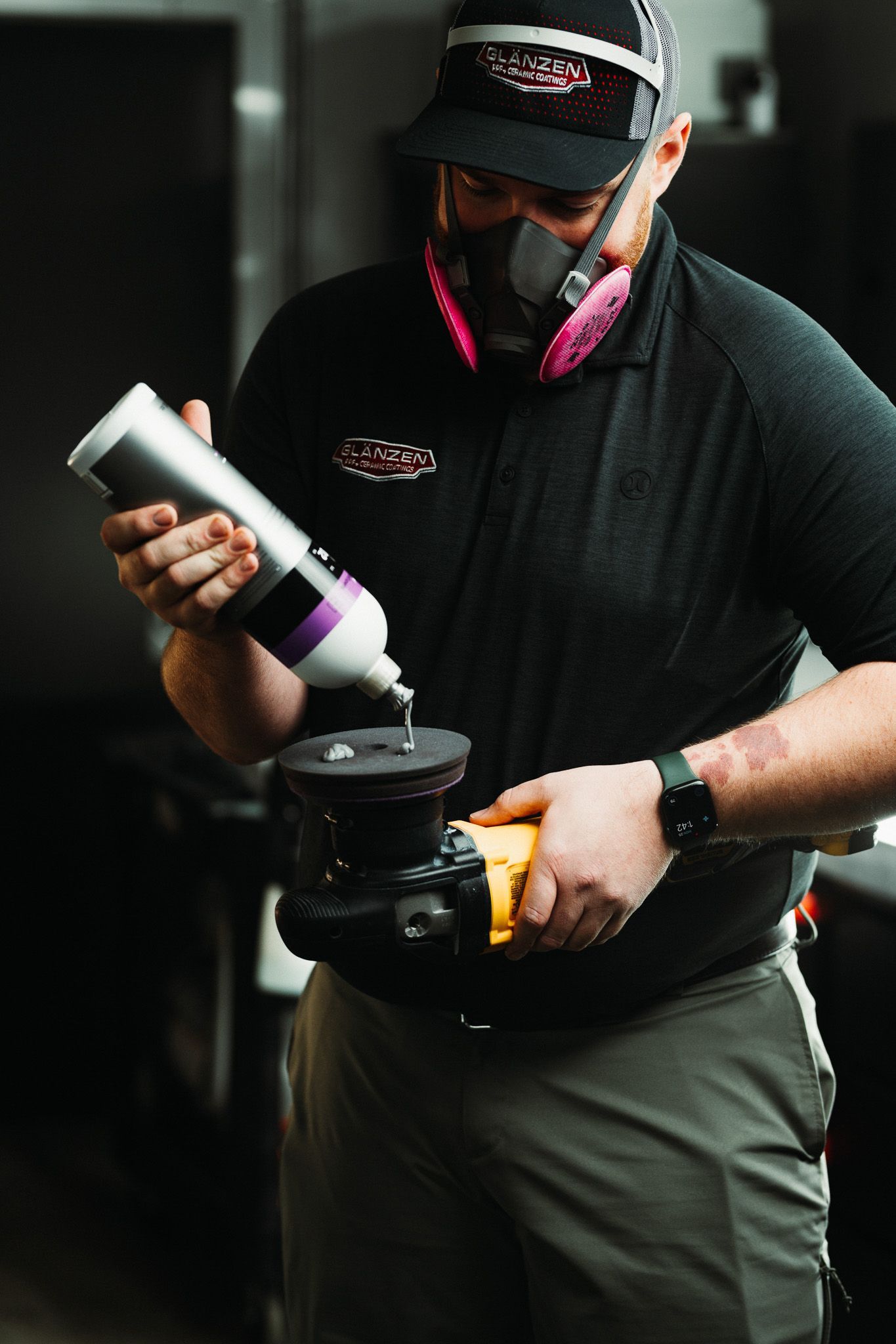 A man wearing a mask is holding a bottle of liquid and polishing a piece of metal.