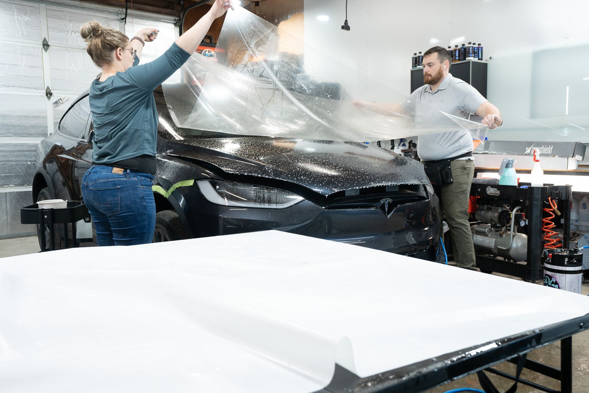 A man and a woman are wrapping a car in a garage.