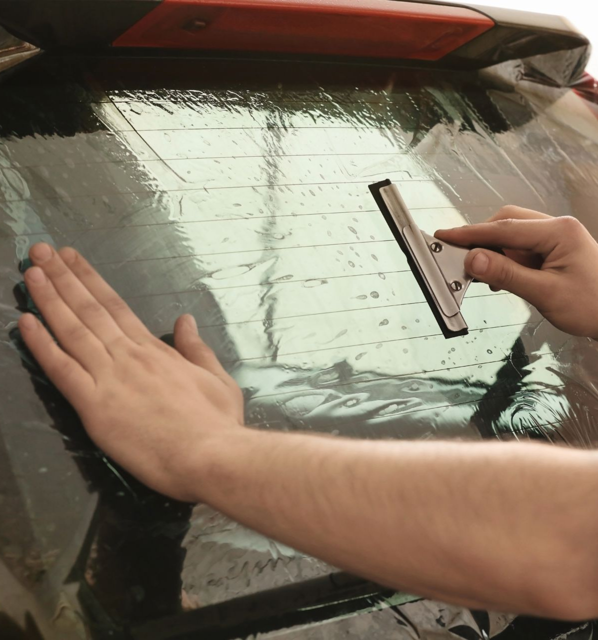 Hands apply a window tint film while using a squeegee on a car rear window.