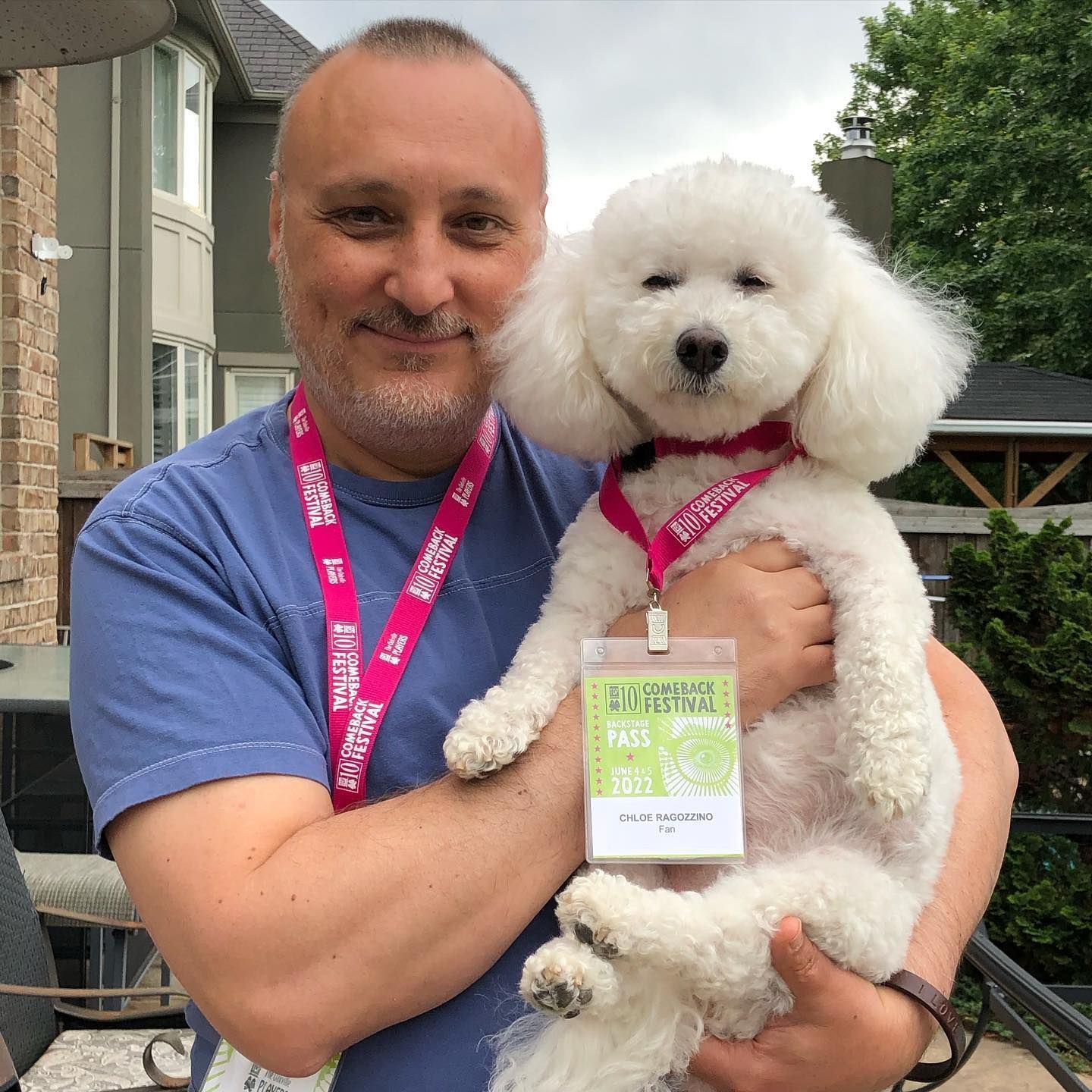 Man holding a small white dog wearing a pink lanyard and badge outdoors in front of houses