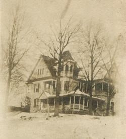 A vintage, multi-story house with a porch, trees, and a snow-covered yard.
