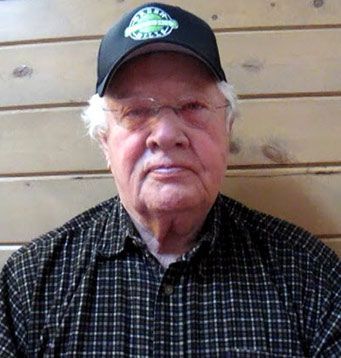 A man wearing a black hat and glasses is sitting in front of a wooden wall.