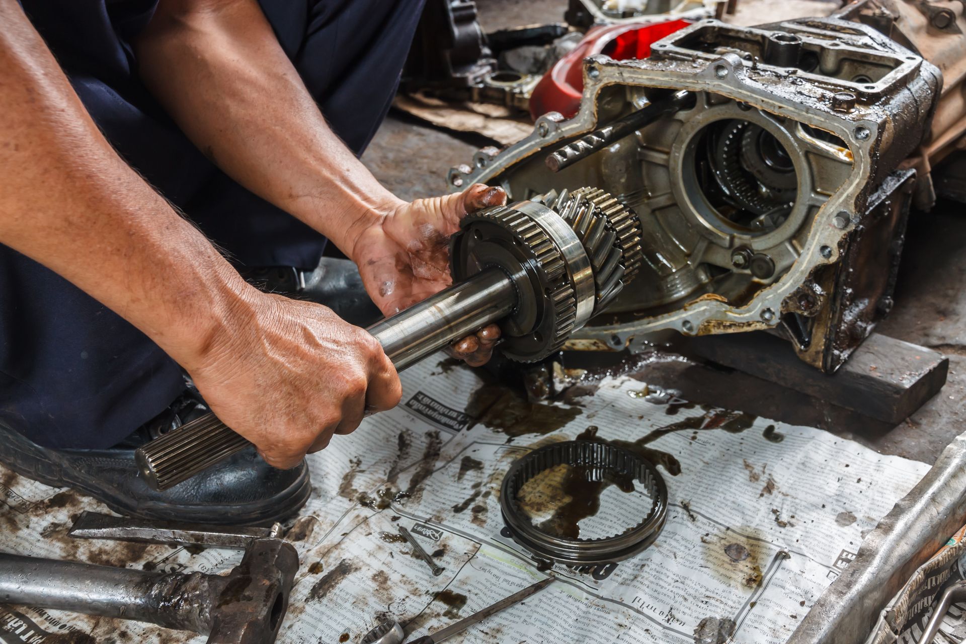 Mechanic working on a transmission, holding gears. Oily workbench, tools, and black shoes are visible.