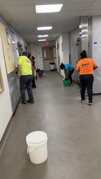 People in safety vests cleaning a hallway. Buckets and cleaning supplies are present.