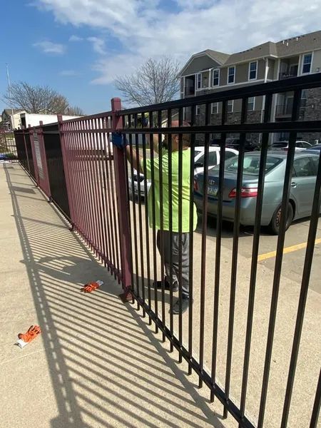 Person painting a black metal fence with a red section. Cars and apartment buildings in background.