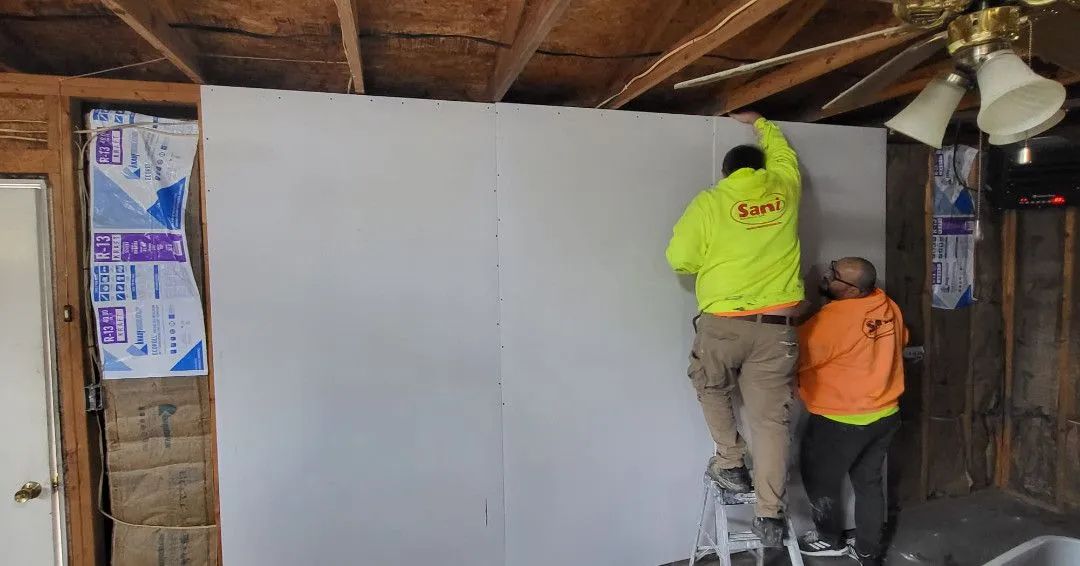 Two workers installing drywall in a room; one on a ladder, the other assisting.