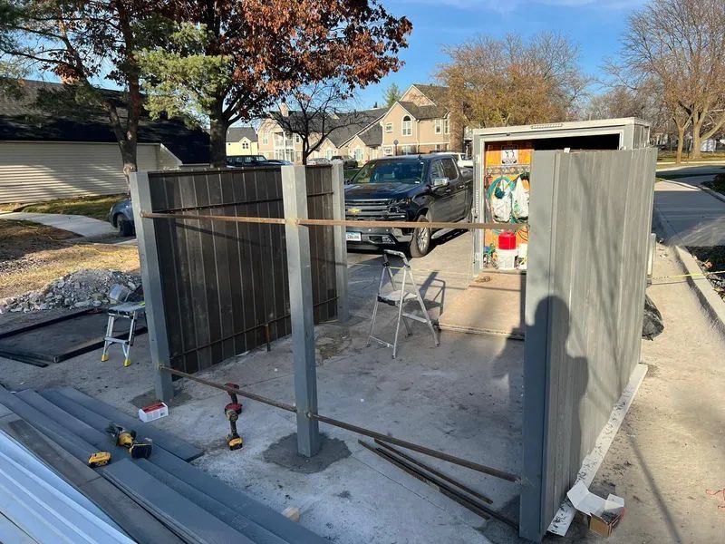 Construction site with a partially built fence; concrete posts, wooden panels. A vehicle is parked nearby.