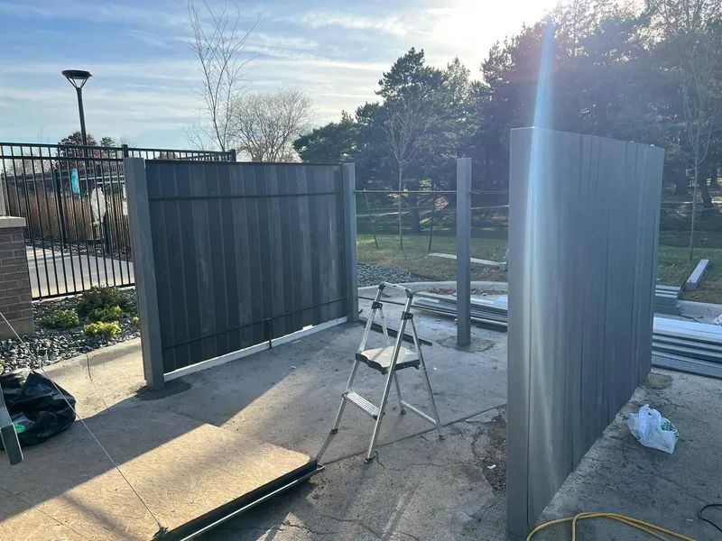 Construction of a privacy fence outdoors with dark gray vertical panels. A stepladder and tools are present.
