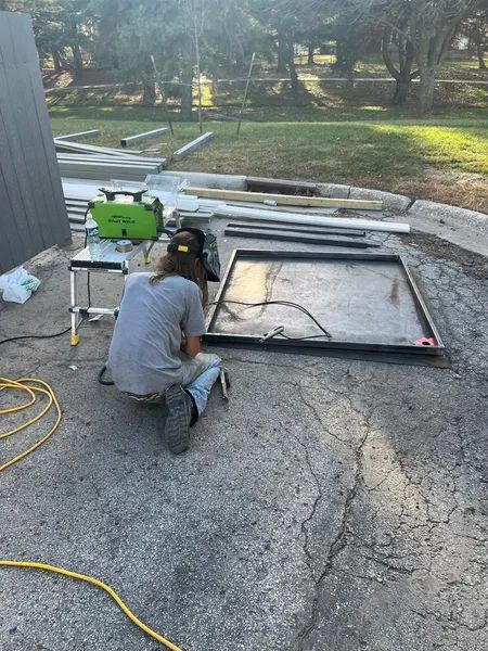 Person welds metal frame outdoors, using welding equipment on a cracked asphalt surface.