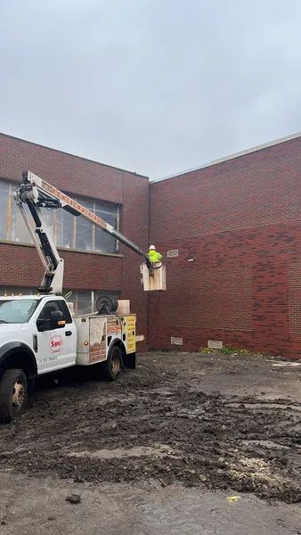 Worker in lift bucket repairing building exterior. Muddy ground. Truck parked by brick wall.