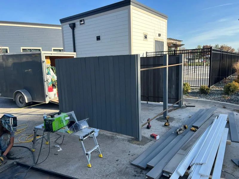 Construction site with a partially built gray privacy fence, tools, and trailer. A person welds.