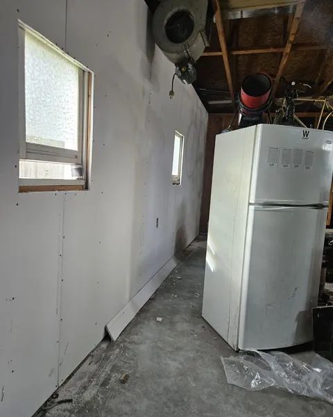 A narrow, unfinished room with drywall walls, two windows, a refrigerator, and a concrete floor.