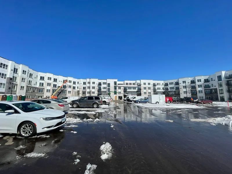 Apartment building with cars parked on a wet, icy lot under a bright blue sky.