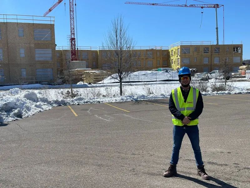 Construction worker in safety vest and hard hat stands in front of unfinished building and snowy ground.