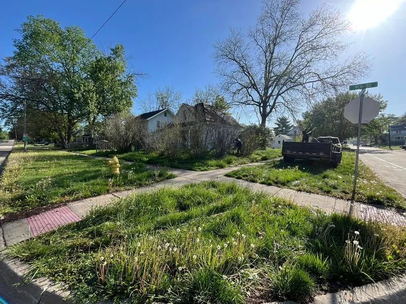 Overgrown lot with a dilapidated house at a street corner on a sunny day. A truck is parked on the side.