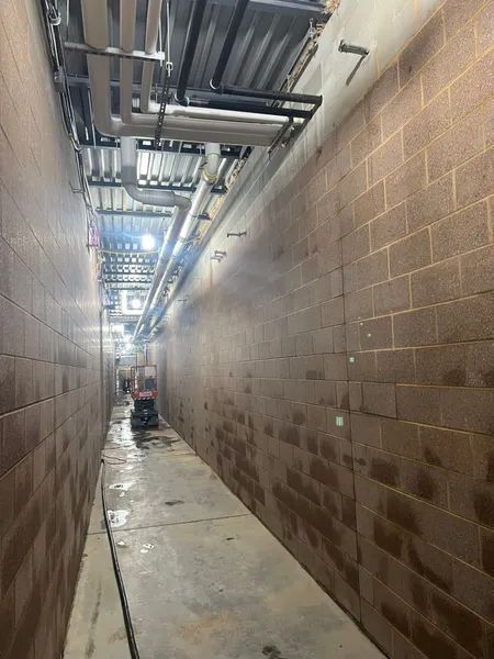 Narrow hallway with concrete block walls, pipes overhead, and a person in the distance.