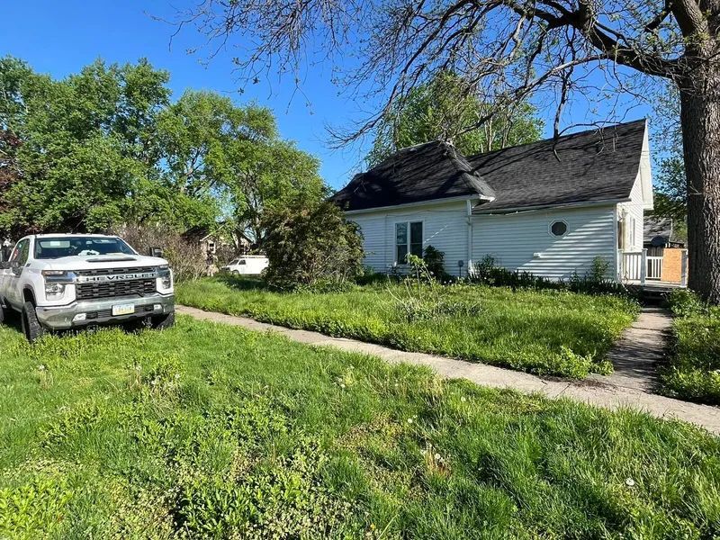 White house with overgrown lawn; white truck parked in front.