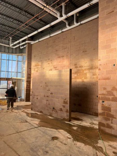 Interior of a building under construction, showing brick walls, water damage, pipes, and a worker walking.