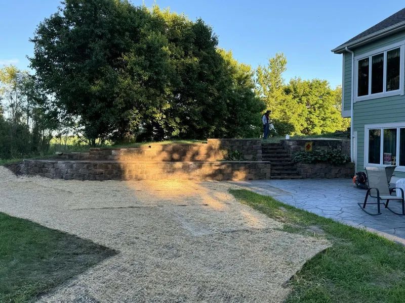 Backyard scene with gravel, retaining wall, stairs, and a house. Green grass and trees are in the background.