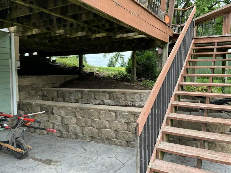 Stone steps and wooden stairs leading up to a deck, under a brown deck with a black handrail.