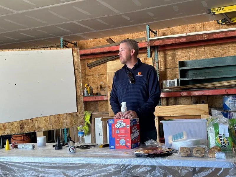 Man stands by a table with snacks and supplies in a workshop. He looks towards the left.