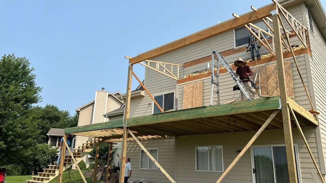 Construction workers building a two-story wooden deck attached to a house. One worker on a ladder.