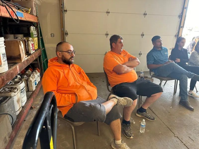 Three men in work attire seated, listening. Garage setting with shelves and open door.