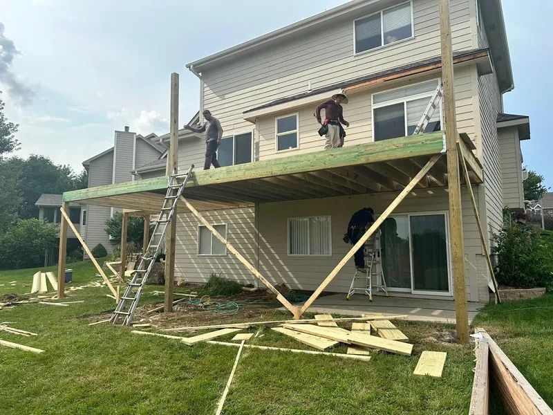 Deck construction: Workers building a deck attached to a two-story beige house; lumber and tools are visible.